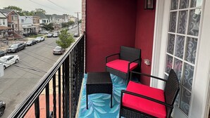 A balcony with two wicker chairs and a table, a red wall, and a view of a street with parked cars. - Cityscape Retreat In Jersey City. Spacious apt for short and long term stay. (Jersey City)