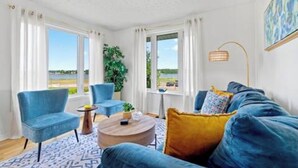 A room with a blue sofa, a round coffee table, and a view of the outdoors through large windows. - Waterview Retreat| Large Deck | Minutes to Downtown  (Charlottetown)