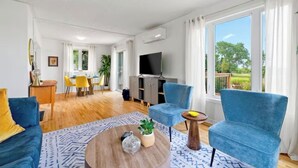 A modern living room with a sofa, armchairs, a coffee table, and a TV. There is a dining area with chairs and a table, and a balcony with a view of greenery. - Waterview Retreat| Large Deck | Minutes to Downtown  (Charlottetown)