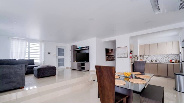 A modern living room with a sofa, ottoman, and a TV on a wooden stand. There is a dining area with a table and chairs, and a kitchen with cabinets and appliances. - Dream House on Isla Verde Beach + Terrace (Carolina)