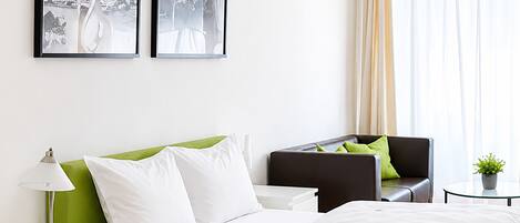 Una habitación de hotel moderna con cama, mesitas de noche, lámpara, silla, una pequeña mesa con una planta y dos cuadros en la pared.