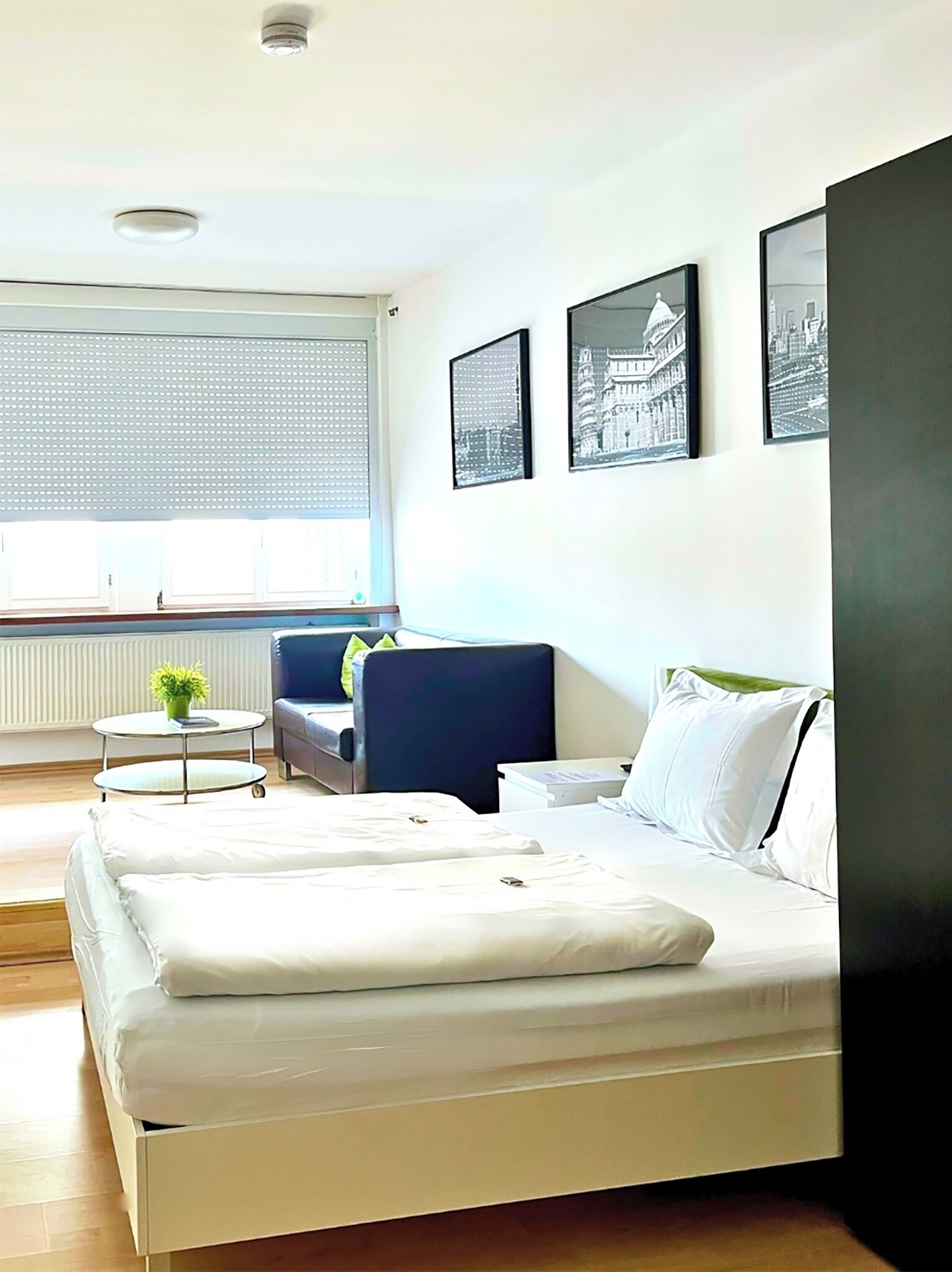 A hotel room with a bed, a chair, a small table, and three framed pictures on the wall.