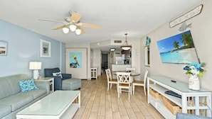 A living room with a sofa, coffee table, and TV stand. There is a dining area with a table and chairs. The room has a ceiling fan and a painting of a beach scene. - Atlantica III PH64- Oceanfront 1BR- Lazy River (Myrtle Beach)