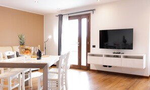 Een moderne woonkamer met een eethoek, een flatscreen televisie bevestigd aan een wit meubelstuk en een schuifpui naar een balkon.