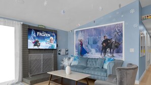 A living room with a grey sofa, a coffee table, a fireplace, a TV displaying 'Frozen', and a large 'Frozen' poster on the wall.