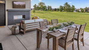 An outdoor patio with a wooden table set for dining, a fireplace, and a TV mounted on the wall. - Welcome to Lucky 13 • Luxurious, private escape on Lake Hills Golf Course. (Billings)