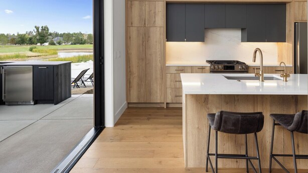 A modern kitchen with wooden cabinets, a central island with bar stools, and a view of a patio area. - Welcome to Lucky 13 • Luxurious, private escape on Lake Hills Golf Course. (Billings)