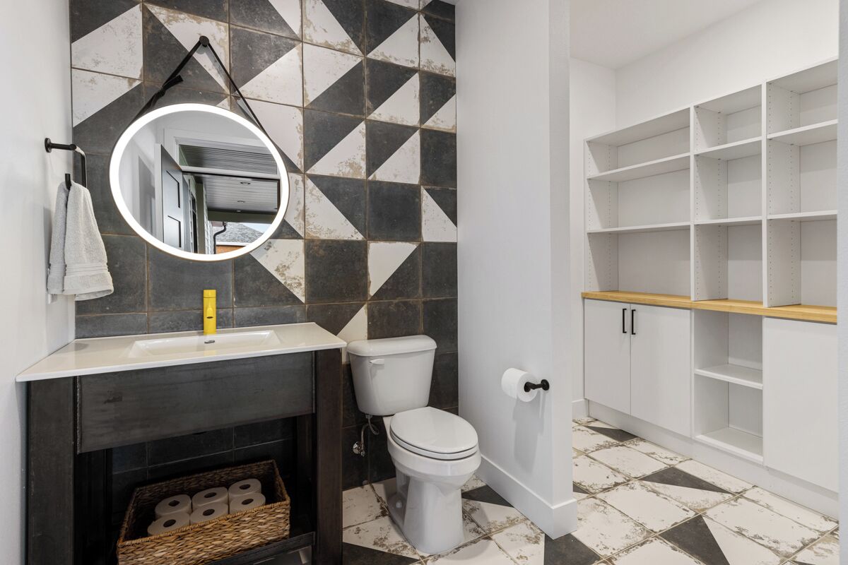Un bany amb paret de rajoles negres i blanques, un vàter blanc, un mirall, un lavabo i un moble prestatge de fusta.