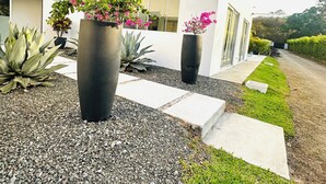 A modern building with a white exterior, large black planters with flowering plants, and a gravel pathway leading to a grassy area. - Kai Guana House (Provincia de Guanacaste)