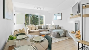 Un salon moderne comprenant un coin repas, un ensemble de canapés, un téléviseur fixé au mur et une grande fenêtre donnant sur la verdure.