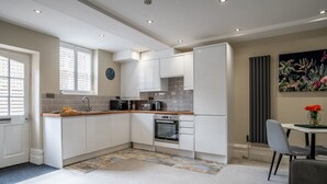 Una cocina moderna con armarios blancos, salpicadero de azulejos y horno integrado. Hay una zona de comedor con mesa y sillas, y un cuadro en la pared.