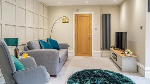 Una sala de estar moderna con un sofá gris, cojines turquesa, una puerta de madera, un televisor y una pequeña mesita de madera con un florero.