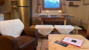 A cozy room with a brown leather sofa, a wooden table with a book and a pink item, a window with a mountain view, a small kitchen area, and a wall-mounted TV. - 50m from ski slopes, 2nd floor, view mountain, balcony, tv, 52m², La Plagne (La Plagne)