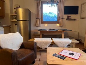 Um quarto acolhedor com um sofá de couro marrom, uma mesa de madeira com um livro e um objeto rosa, uma janela com vista para as montanhas, uma pequena área de cozinha e uma TV presa à parede.