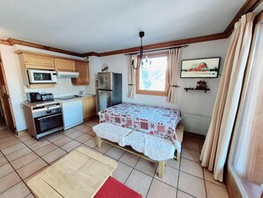 Quarto compacto com cozinha compacta, uma cama com colcha estampada, uma mesa pequena e uma janela com cortinas.
