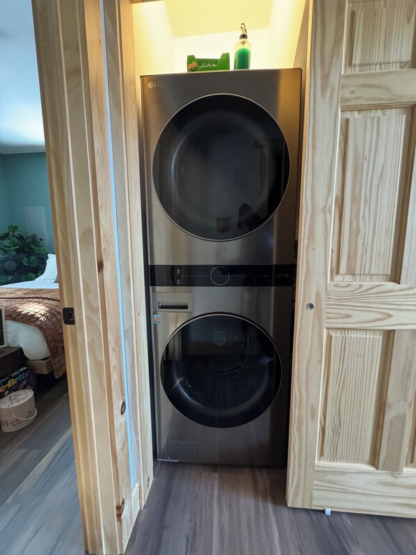 A compact, modern laundry room with two large washing machines, a wooden door, and a small shelf with bottles. - Fall Foliage at Modern Artsy Lux Cabin w/BBQ, Pond, Fire pit near Hiking + Shops (Narrowsburg)