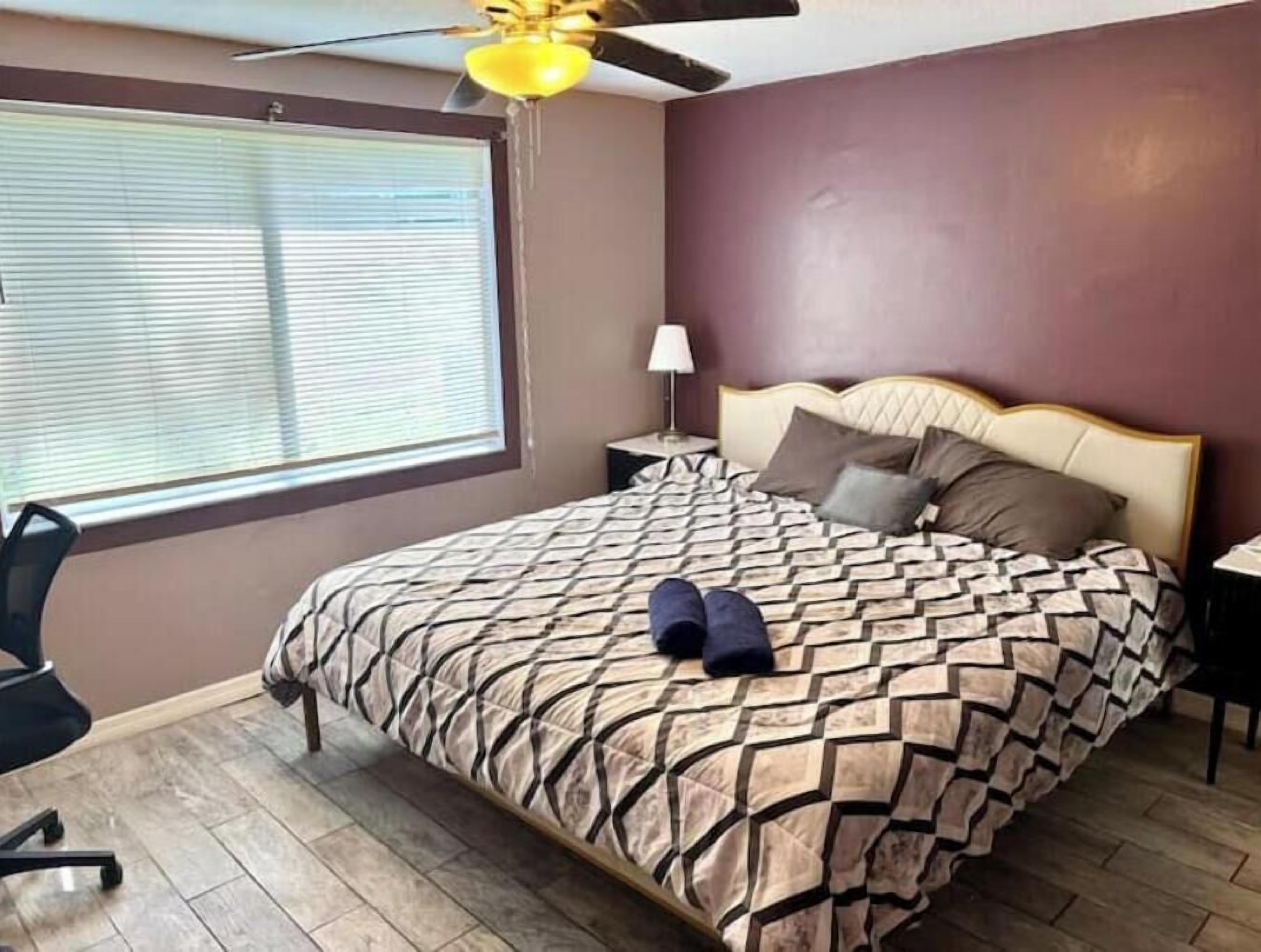 Un dormitorio con cama, ventilador de techo, mesita de noche, lámpara y ventana con persianas.