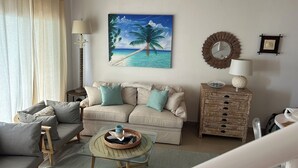 A living room with a beige sofa, a glass coffee table, a wooden side table, and a painting of a beach scene.