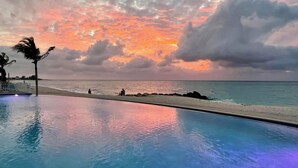 A poolside view with a sunset over the ocean, palm trees, and a beach.