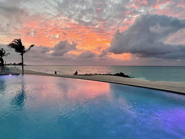Vista de la piscina con un atardecer sobre el océano, palmeras y una playa.