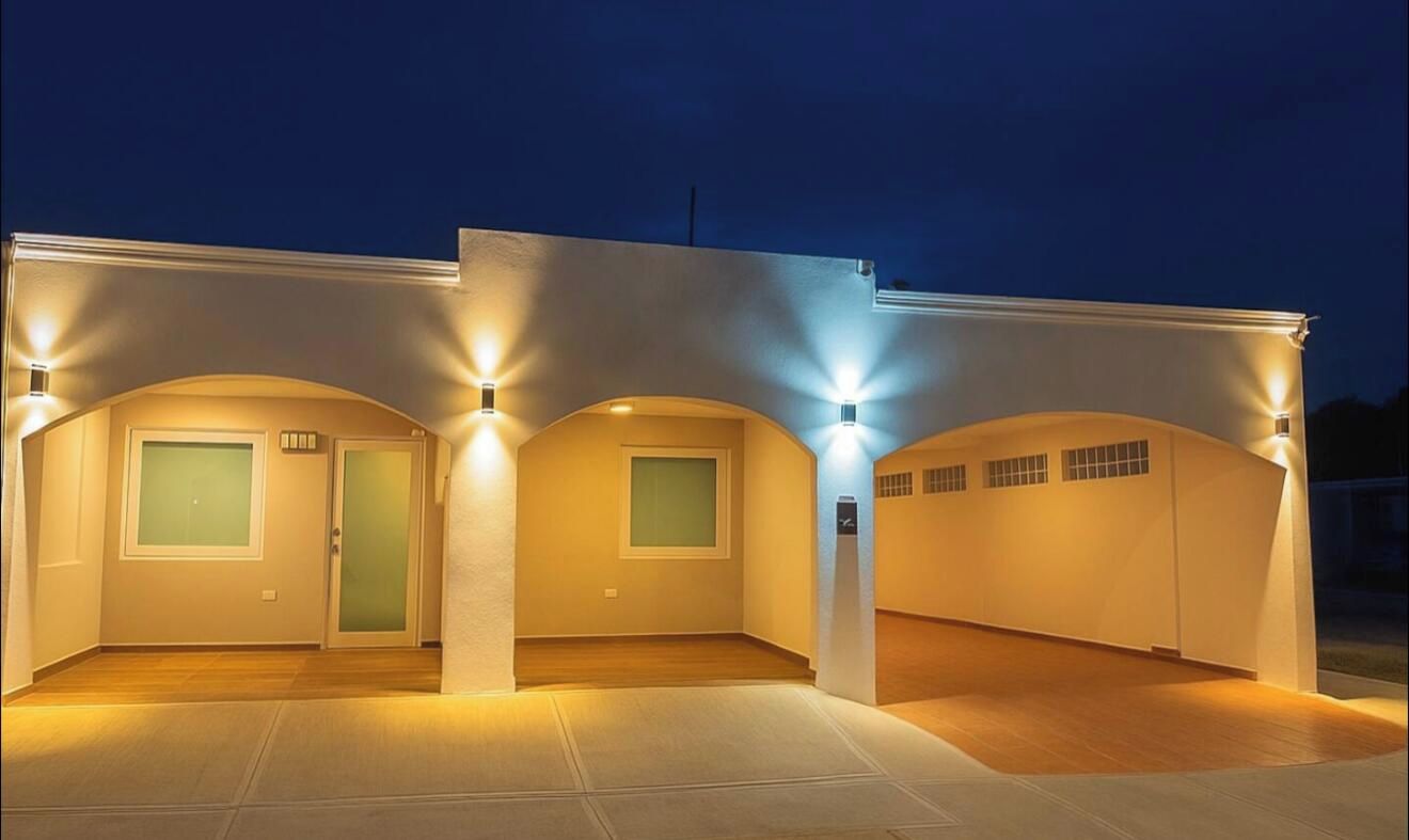 Un edificio moderno con tres ventanas iluminadas y una puerta, que presenta un techo plano y un exterior de color claro.