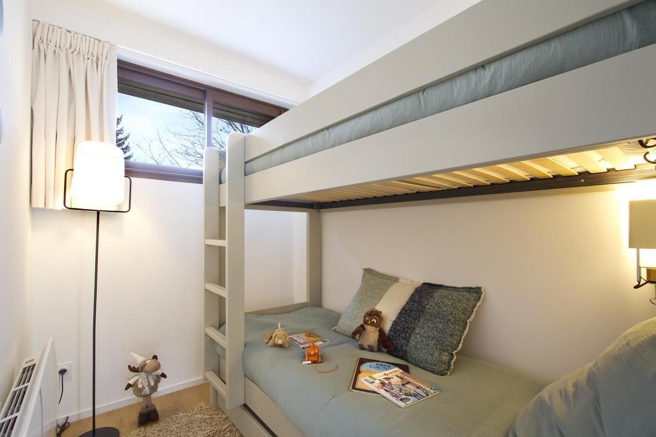 A bunk bed room with a window, a lamp, a stuffed toy, and a magazine on the bed.