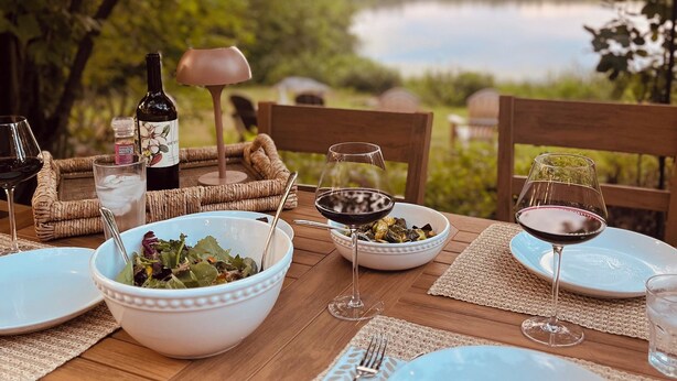 An outdoor dining table set with plates, bowls, wine glasses, and cutlery, overlooking a lake. - Serene Waterfront Retreat on 2+ Acres with 360-degree Water Views & Private Dock (Center Harbor)