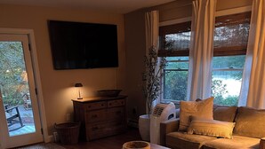 A cozy living room with a sofa, a coffee table, and a TV. There is a window with curtains and a view of greenery outside. - Serene Waterfront Retreat on 2+ Acres with 360-degree Water Views & Private Dock (Center Harbor)