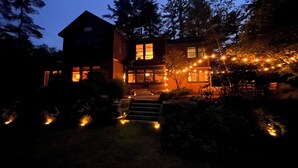 A two-story house with a front porch, illuminated by string lights and surrounded by trees. - Serene Waterfront Retreat on 2+ Acres with 360-degree Water Views & Private Dock (Center Harbor)