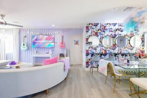 A modern living room with a curved white sofa, a glass coffee table, and a pink chair. There is a wall with a TV and a shelf with various items.