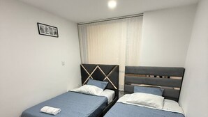 A hotel room with two beds, each with a blue bedspread and white pillows. There is a framed picture on the wall and a window with curtains. - Apartment for 8 guests in the center of Pasto 3 bedrooms 2 bathrooms (Pasto)