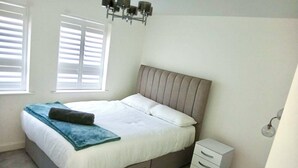 A bedroom with a bed, a nightstand, a dresser, and a window with blinds. - Grand 4-bed House in Ebbsfleet Valley, Garden & Parking Space (Swanscombe)