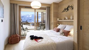 A cozy bedroom with a large bed, a wooden headboard, a view of mountains, and a balcony with chairs.