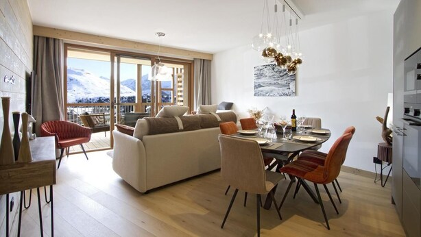 A spacious living room with a large sofa, dining table, and chairs. There is a balcony with a view of snow-capped mountains.