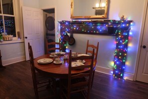 A dining room with a round wooden table set for four, chairs, a fireplace with colorful lights, a mirror above the fireplace, and a door leading to another room.