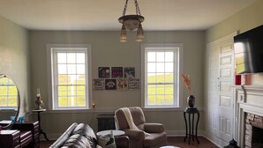 A living room with a leather sofa, a coffee table, and a fireplace. There are two windows with curtains, a wall-mounted TV, and a chandelier. - Historic Farm Stay at The Hager Hideaway (Harrodsburg)