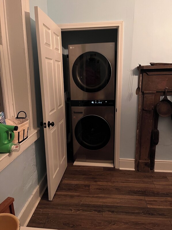 Une buanderie équipée d’un lave-linge et d’un sèche-linge à double porte, d’une table en bois sur laquelle se trouve un pot, et d’un récipient vert posé sur le sol.