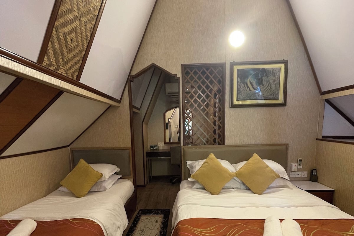 A hotel room with two beds, a wooden floor, a decorative wall panel, and a small table with a lamp.