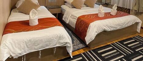 Habitación de hotel con dos camas, un candelabro, un ventanal grande con cortinas y una alfombra estampada.