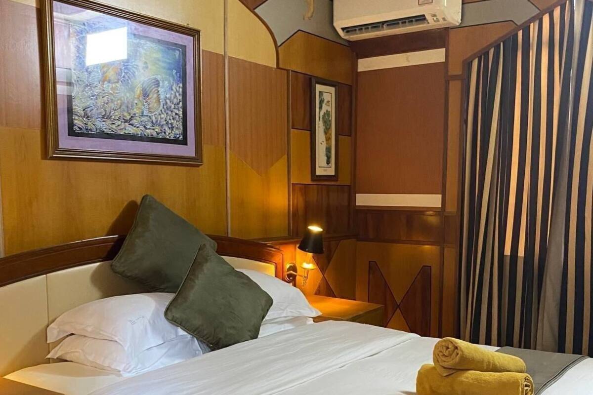 A hotel room with a bed, pillows, a nightstand, a lamp, a framed artwork, an air conditioning unit, and a striped curtain.