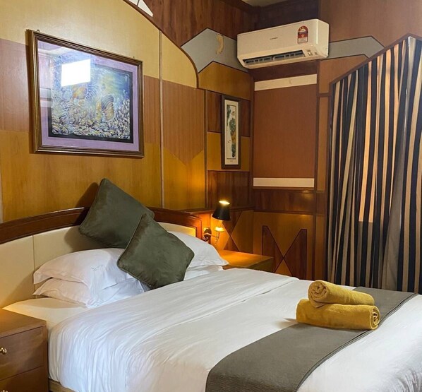Habitación de hotel con cama, almohadas, mesita de noche, lámpara, una obra de arte enmarcada, equipo de aire acondicionado y cortina a rayas.