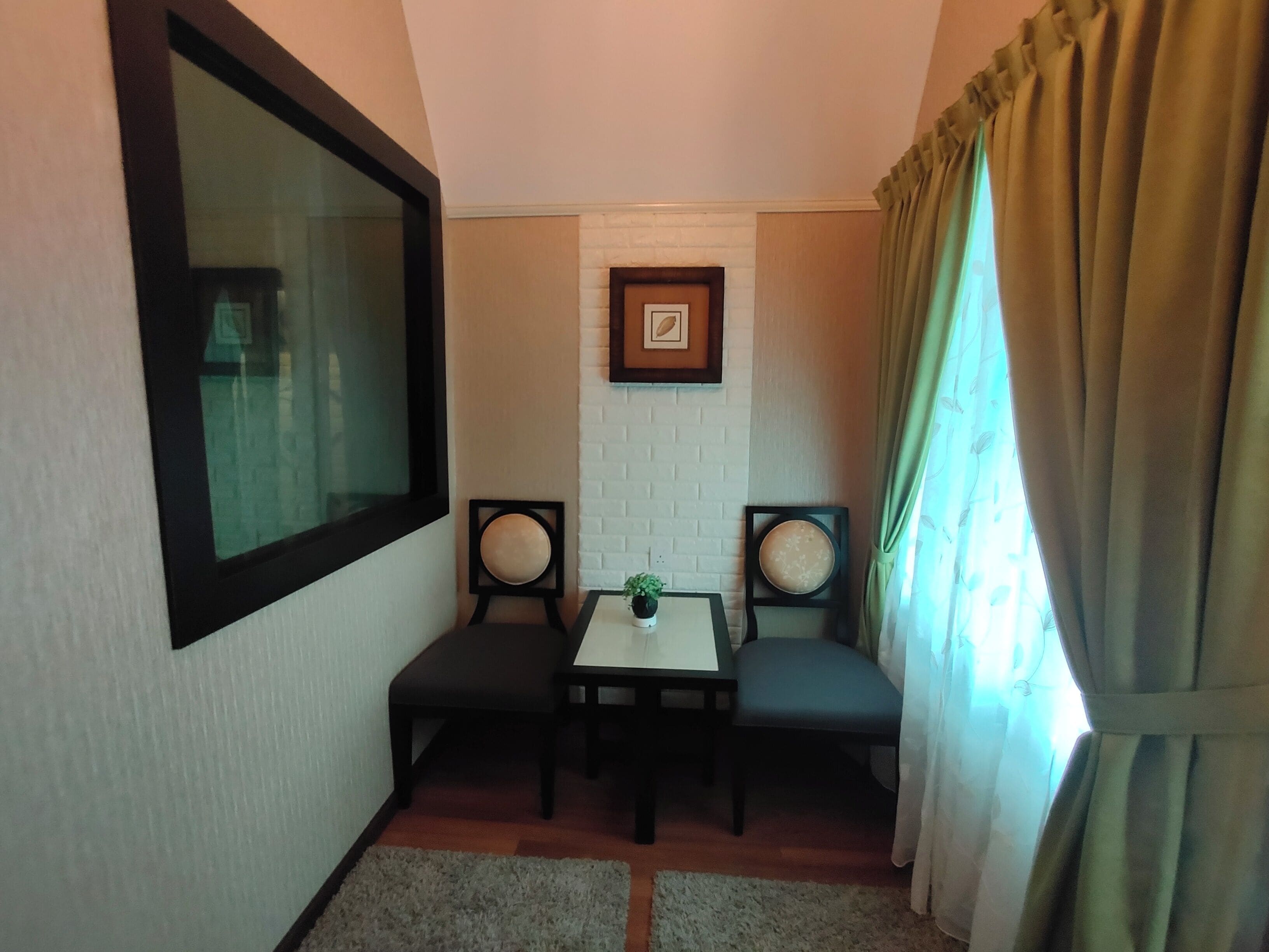 A small, cozy room with a white brick wall, a framed picture, a small table, two chairs, and a window with curtains.