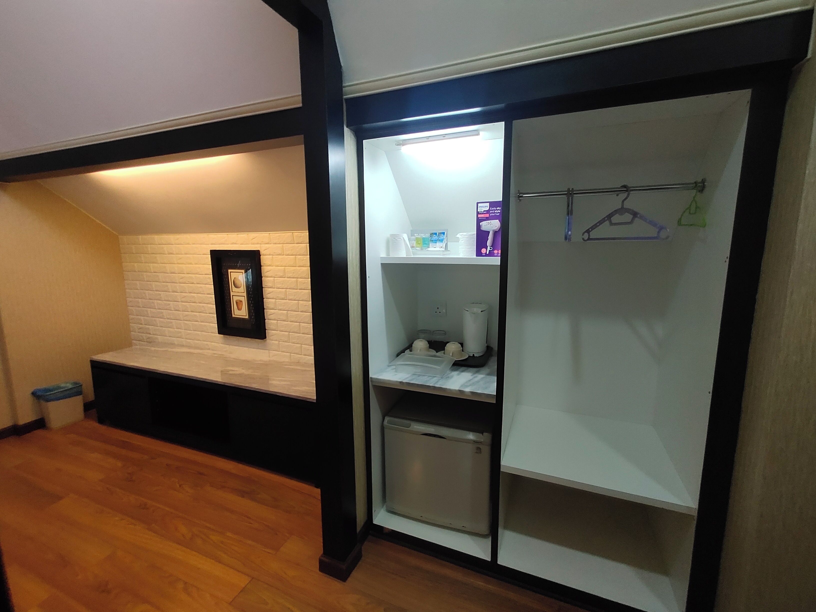 A compact room with a wooden floor, a white brick wall, a framed picture, a small shelf with toiletries, and a white wardrobe with a hanging rod.