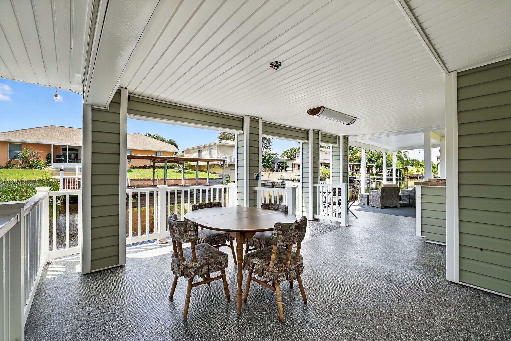 A covered patio with a round table and chairs, a view of a residential area, and a pool in the background.