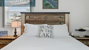 A bedroom with a wooden headboard, a pillow with "JUMP IN THE LAKE" text, a framed painting of a road, and a sign that says "GONE FISHING". - Lake Okeechobee Fishing Retreat (Okeechobee)