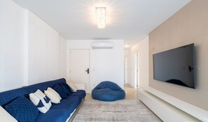 Una sala de estar moderna con un sofá azul, un sillón de saco, un televisor grande y un equipo de aire acondicionado.