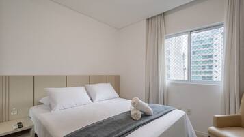 A hotel room with a bed, a chair, a window with curtains, and a view of buildings outside.