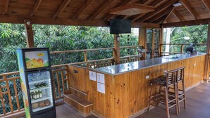 A wooden bar area with a refrigerator stocked with beverages, a wooden counter, and a view of lush greenery.
