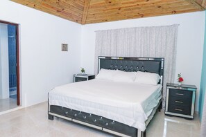 Une chambre à coucher comprenant un lit, deux tables de chevet, une commode, un plafond en bois, une fenêtre avec des rideaux et une porte.