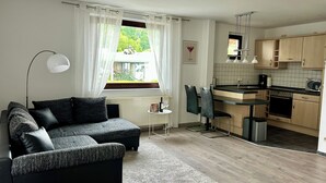 Un salon moderne avec un canapé et un coin repas. On y trouve une fenêtre avec des rideaux, une petite table et une cuisine dotée de meubles en bois et d’appareils électroménagers.